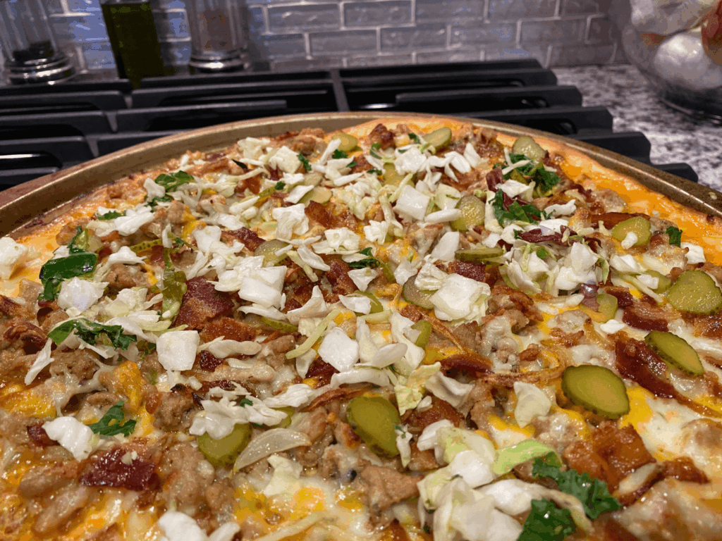 Photo of cheeseburger pizza out of the oven with lettuce, pickles, bacon and ground beef on top