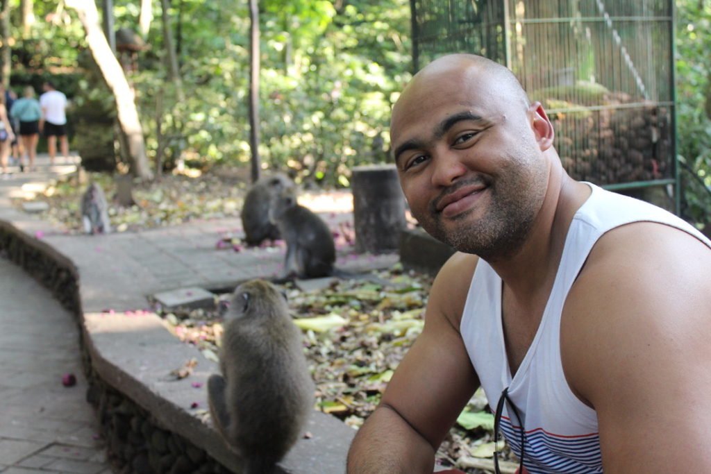 a photo of a man sitting in front of 4 monkeys eating purple potatoes at the Sacred Monkey Forest Sanctuary