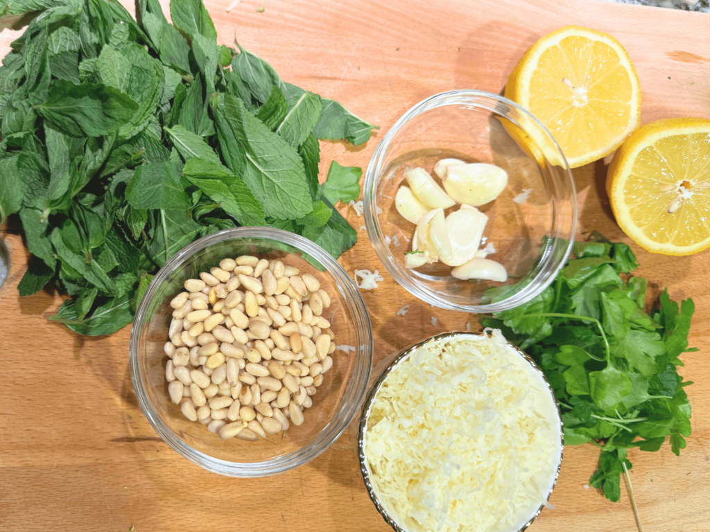 Photo of fresh ingredients needed to make a mint sauce on a wooden cutting board. Parsley, Mint, pine nuts, garlic, parmesan cheese and a lemon cut in half.