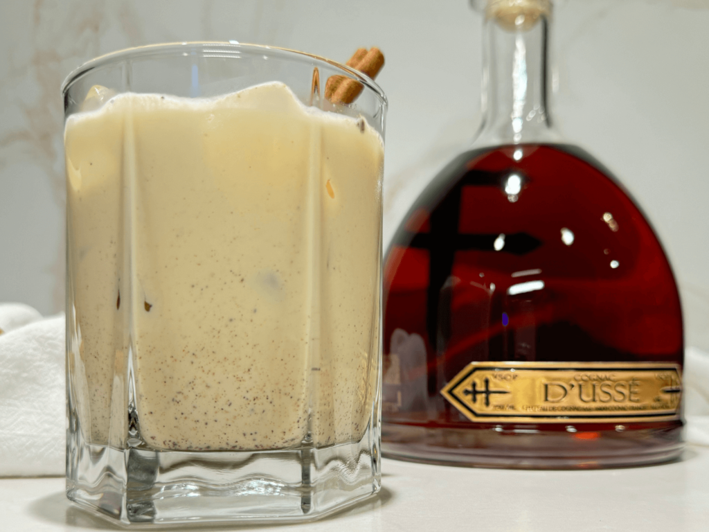 Photo of Homemade Spiked Eggnog in a glass next to a bottle of dusse cognac