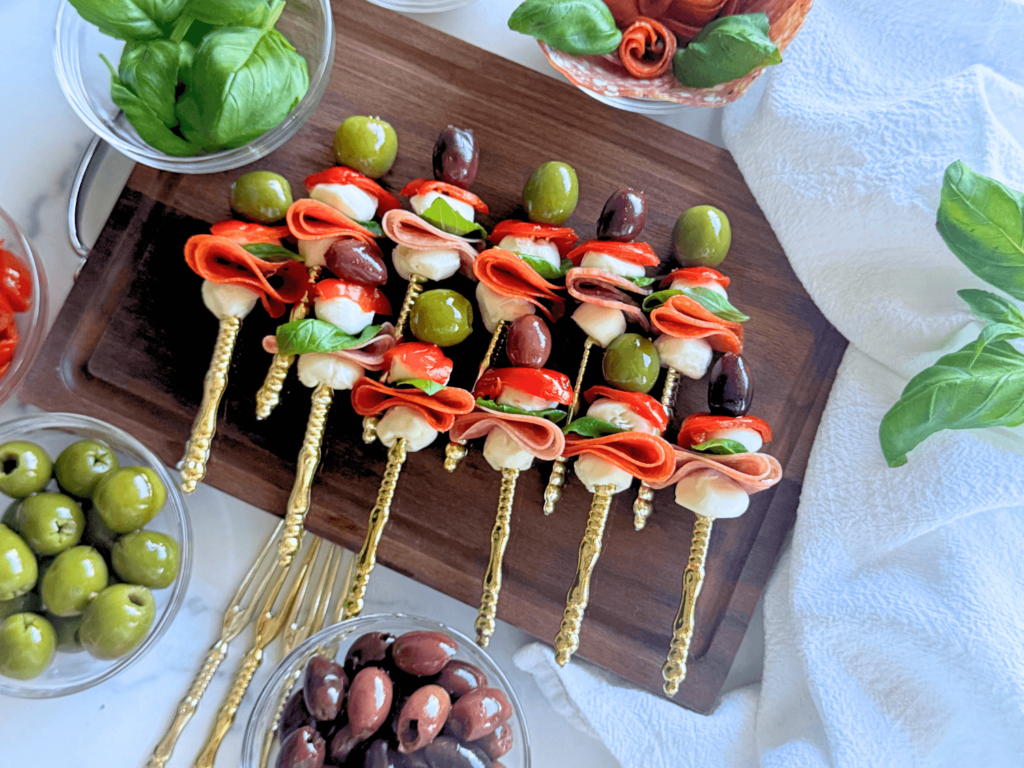 up close photo of antipasto skewers on a wooden cutting board with olives, pepperoni, mozzarella, sweet hot peppers, salami, and basil on a gold fork