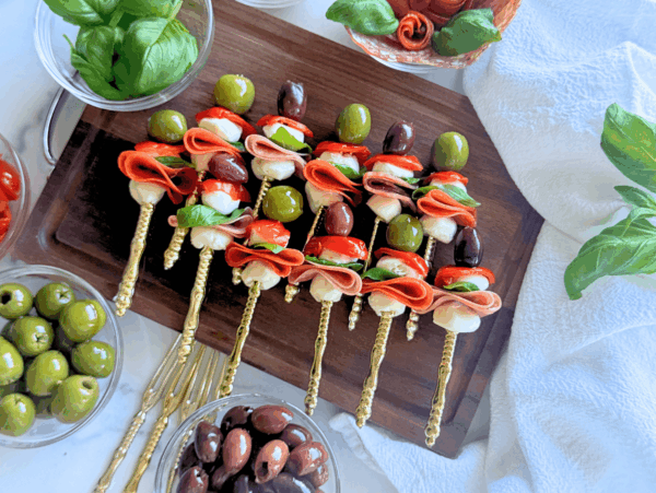 up close photo of antipasto skewers on a wooden cutting board with olives, pepperoni, mozzarella, sweet hot peppers, salami, and basil on a gold fork