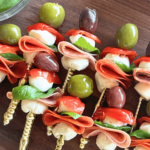 up close photo of antipasto skewers on a wooden cutting board with olives, pepperoni, mozzarella, sweet hot peppers, salami, and basil on a gold fork
