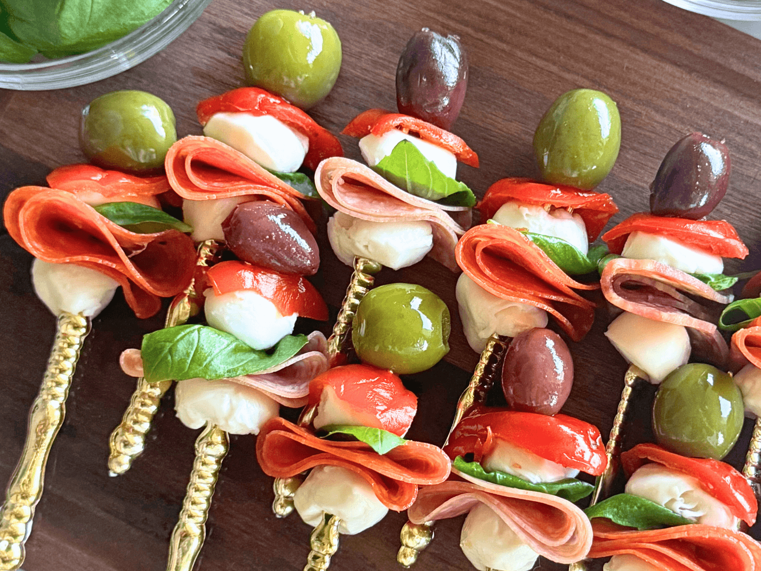 up close photo of antipasto skewers on a wooden cutting board with olives, pepperoni, mozzarella, sweet hot peppers, salami, and basil on a gold fork