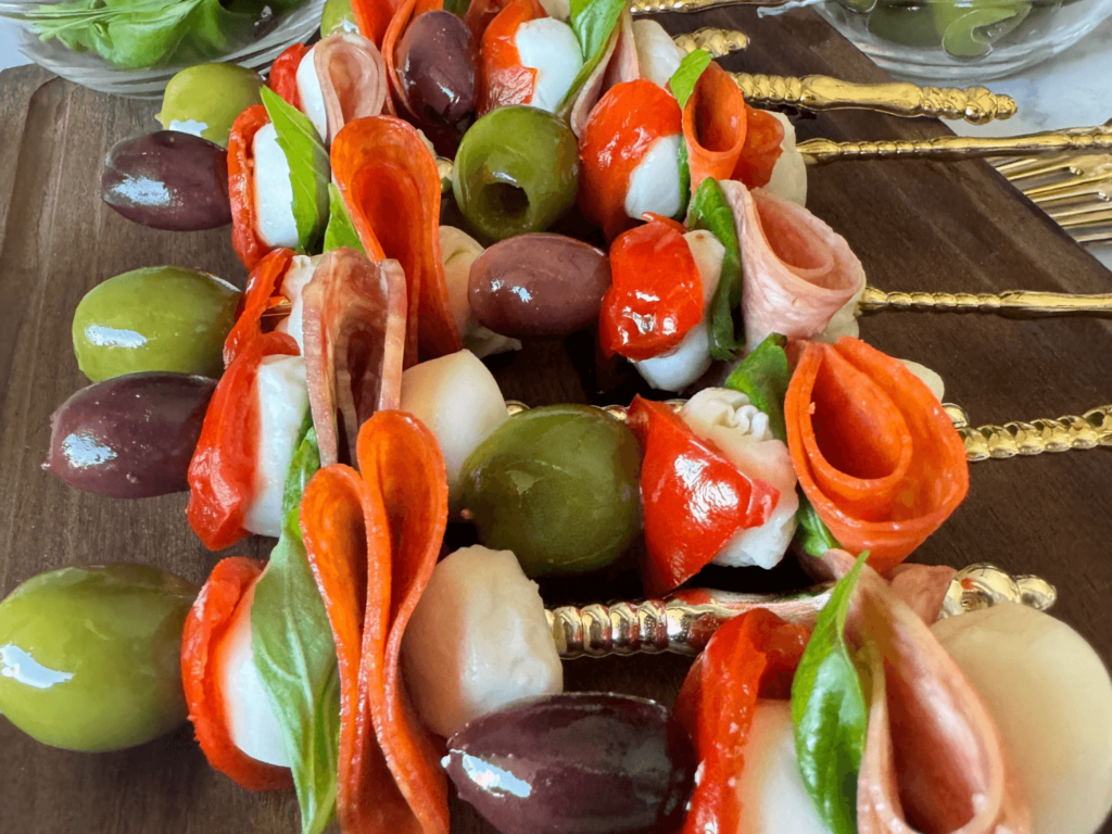 up close photo of antipasto skewers on a wooden cutting board with olives, pepperoni, mozzarella, sweet hot peppers, salami, and basil on a gold fork