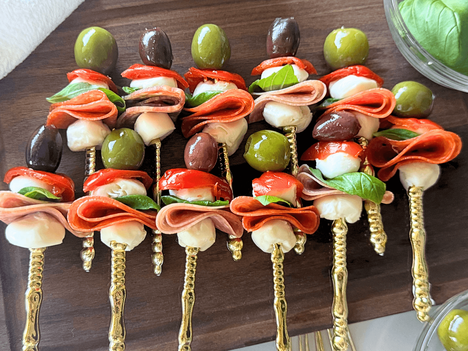 up close photo of antipasto skewers on a wooden cutting board with olives, pepperoni, mozzarella, sweet hot peppers, salami, and basil on a gold fork