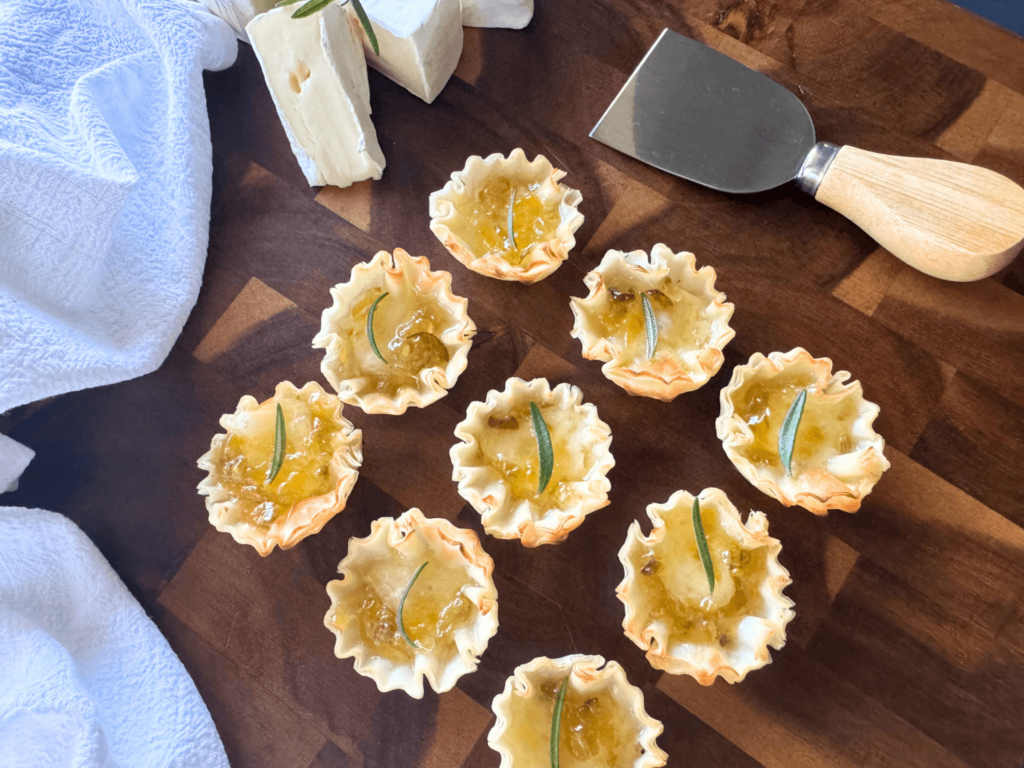 Close up photo of phyllo shells, mini baked brie bites with peach pepper jelly on a wooden cutting board
