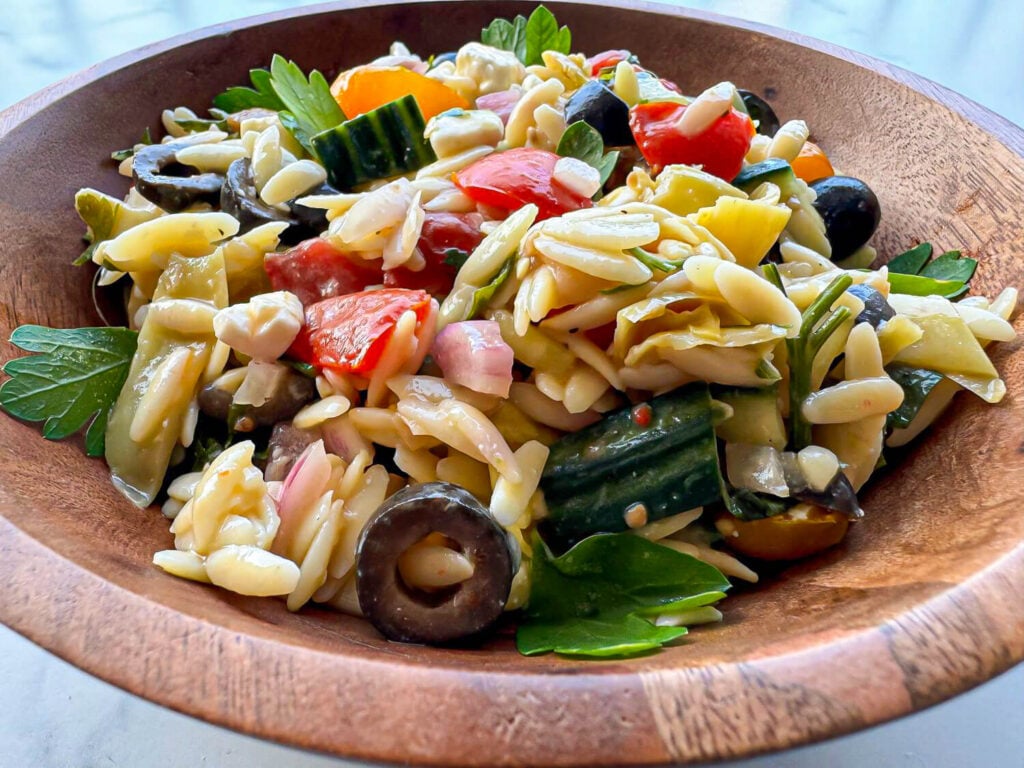 photo of an orzo pasta salad in a wooden bowl. The pasta salad has cooked orzo pasta, chopped parsley, sliced black olives, colorful tomatoes, chopped cucumbers, red onions, and seasonings