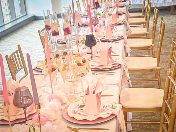 View of tablescape. Table has white spandex table cloth with pink charger plates, cream plates, floral napkins and pink candles