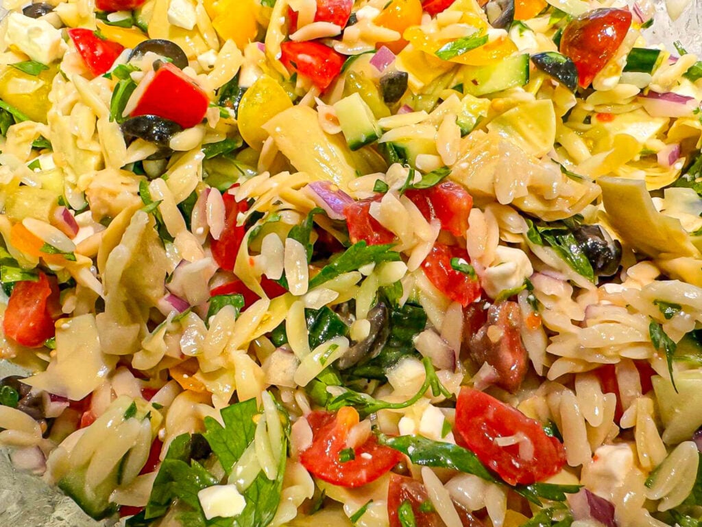 a photo of fresh ingredients in a glass bowl for orzo pasta salad. Chopped artichokes, cherry tomatoes, red onions, olives, cucumbers, feta cheese