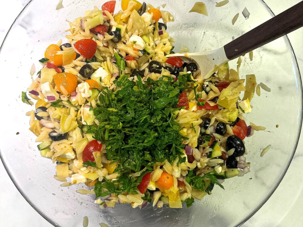 a photo of fresh ingredients in a glass bowl for orzo pasta salad. Chopped artichokes, cherry tomatoes, red onions, olives, cucumbers, feta cheese