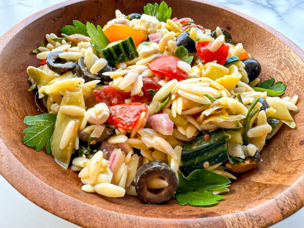 photo of an orzo pasta salad in a wooden bowl. The pasta salad has cooked orzo pasta, chopped parsley, sliced black olives, colorful tomatoes, chopped cucumbers, red onions, and seasonings