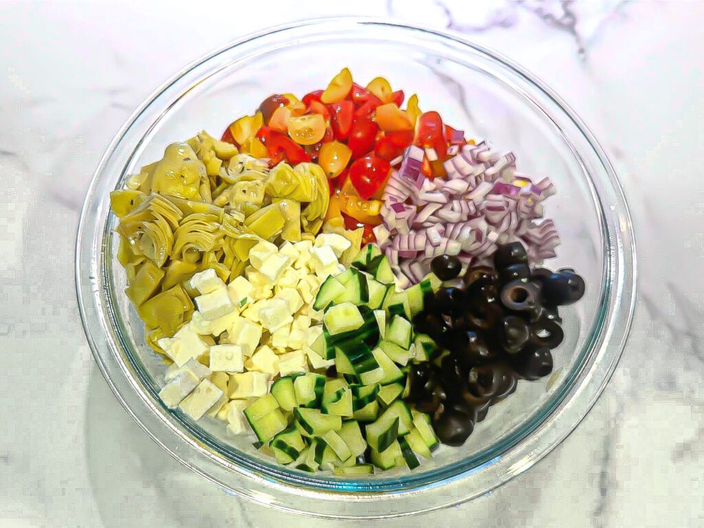 a photo of fresh ingredients in a glass bowl for orzo pasta salad. Chopped artichokes, cherry tomatoes, red onions, olives, cucumbers, feta cheese