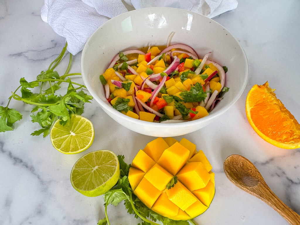 Photo of mango salsa in a white bowl surround by mangos, an orange slice, cilantro and limes