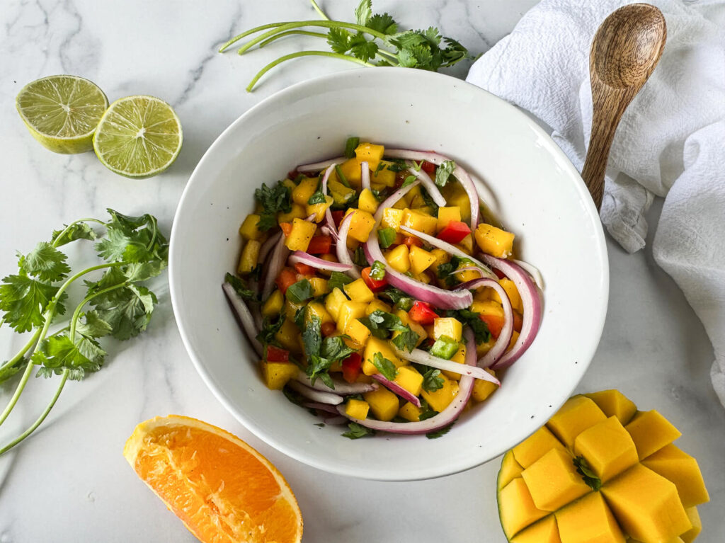 Photo of mango salsa in a white bowl surround by mangos, orange slice, cilantro and limes