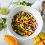 Photo of mango salsa in a white bowl surround by mangos, orange slice, cilantro and limes