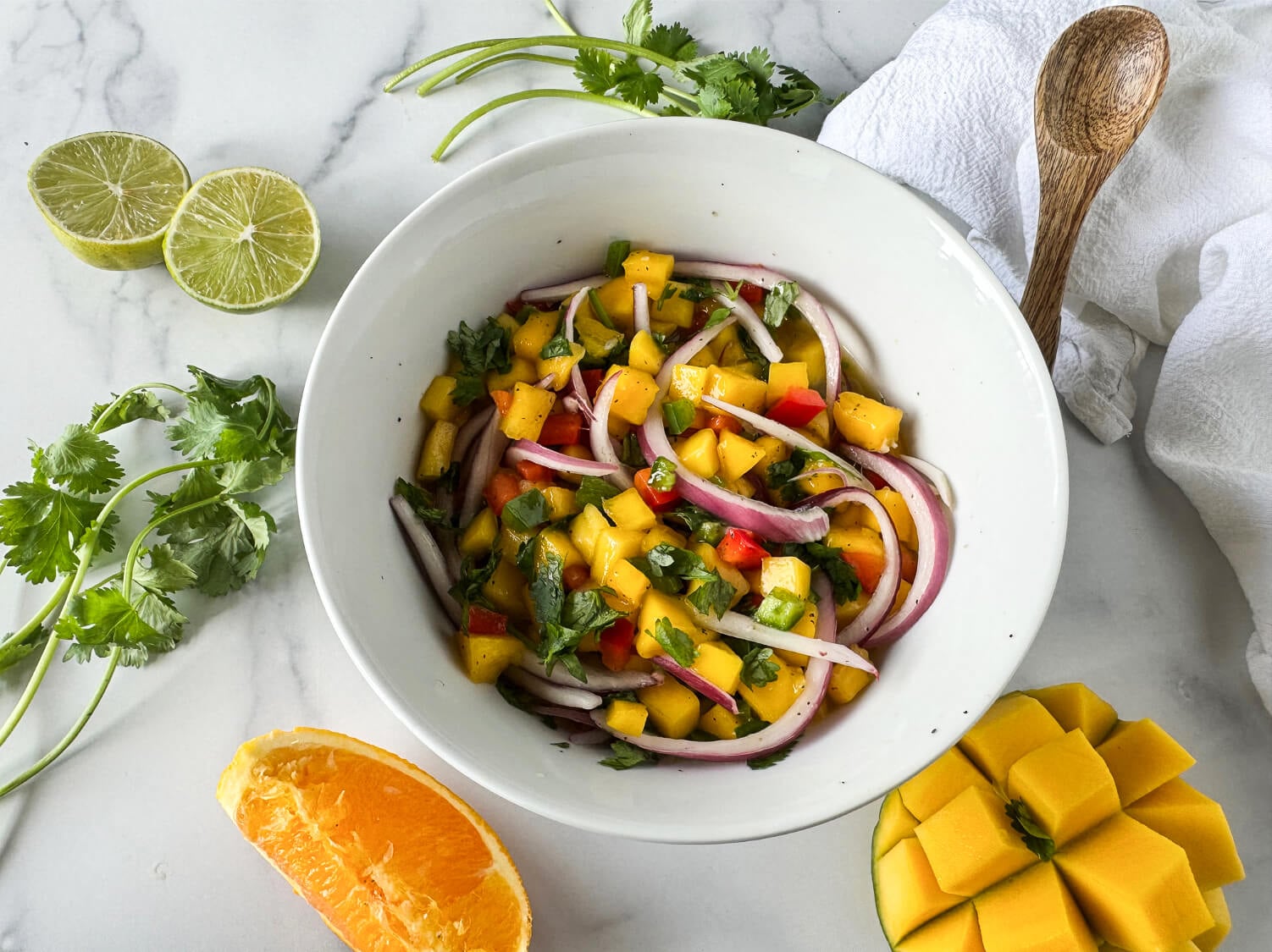 Photo of mango salsa in a white bowl surround by mangos, orange slice, cilantro and limes