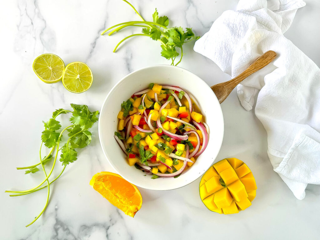 Photo of mango salsa in a white bowl surround by mangos, an orange slice, cilantro and limes