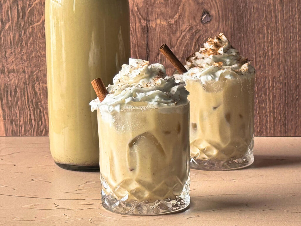 photo of two glasses of dairy-free eggnog topped with dairy-free whipped cream next to a bottle for storage