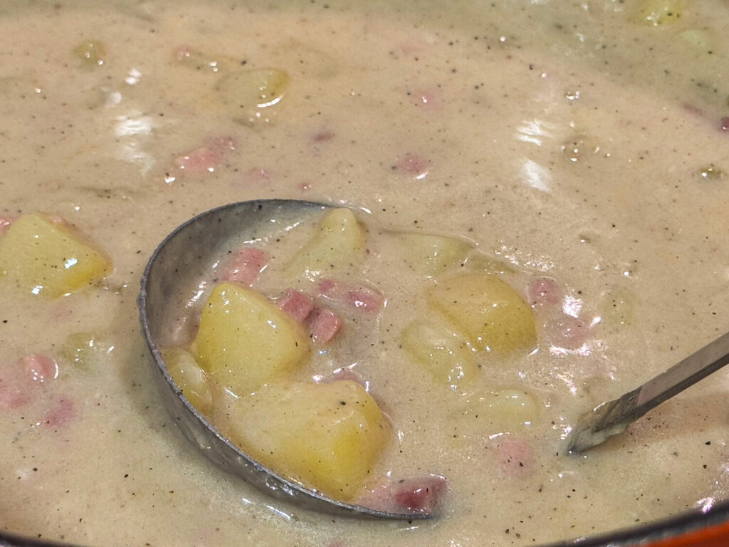 Close up photo of a silver soup spoon scooping ham and potato soup out of the pot.