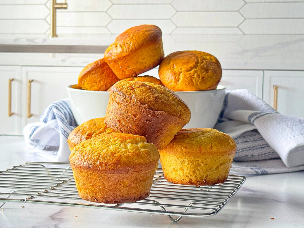photo of Cornbread Muffins on a cooling tray and some stacked in a white bowl in the background