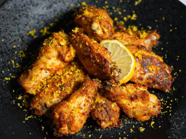 Photo of Air Fryer Lemon Pepper Wings on a black plate with a lemon wedge in the middle and lemon zest