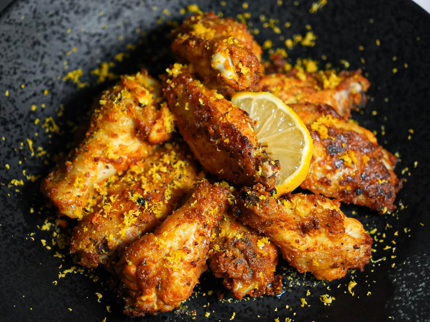 Photo of Air Fryer Lemon Pepper Wings on a black plate with a lemon wedge in the middle and lemon zest