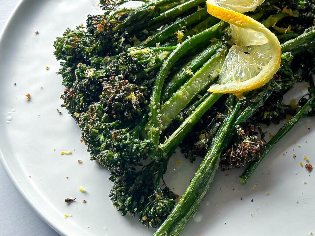 Close up photo of cooked broccolini on a white plate with a lemon twist