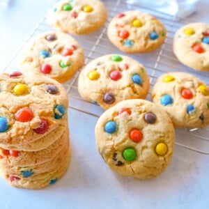 photo of a stack of M&M cookies next to a wire rack tray full of more M&M cookies