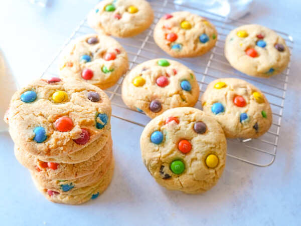 photo of a stack of M&M cookies next to a wire rack tray full of more M&M cookies