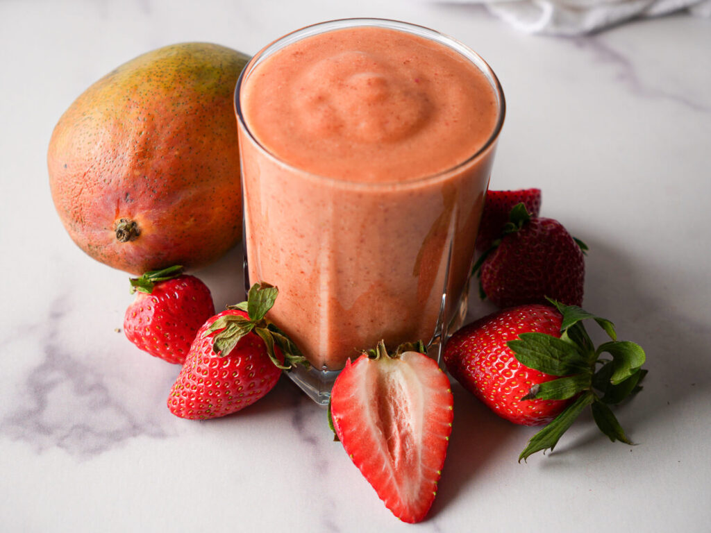 side view of one strawberry mango smoothie surrounded by strawberries and a mango