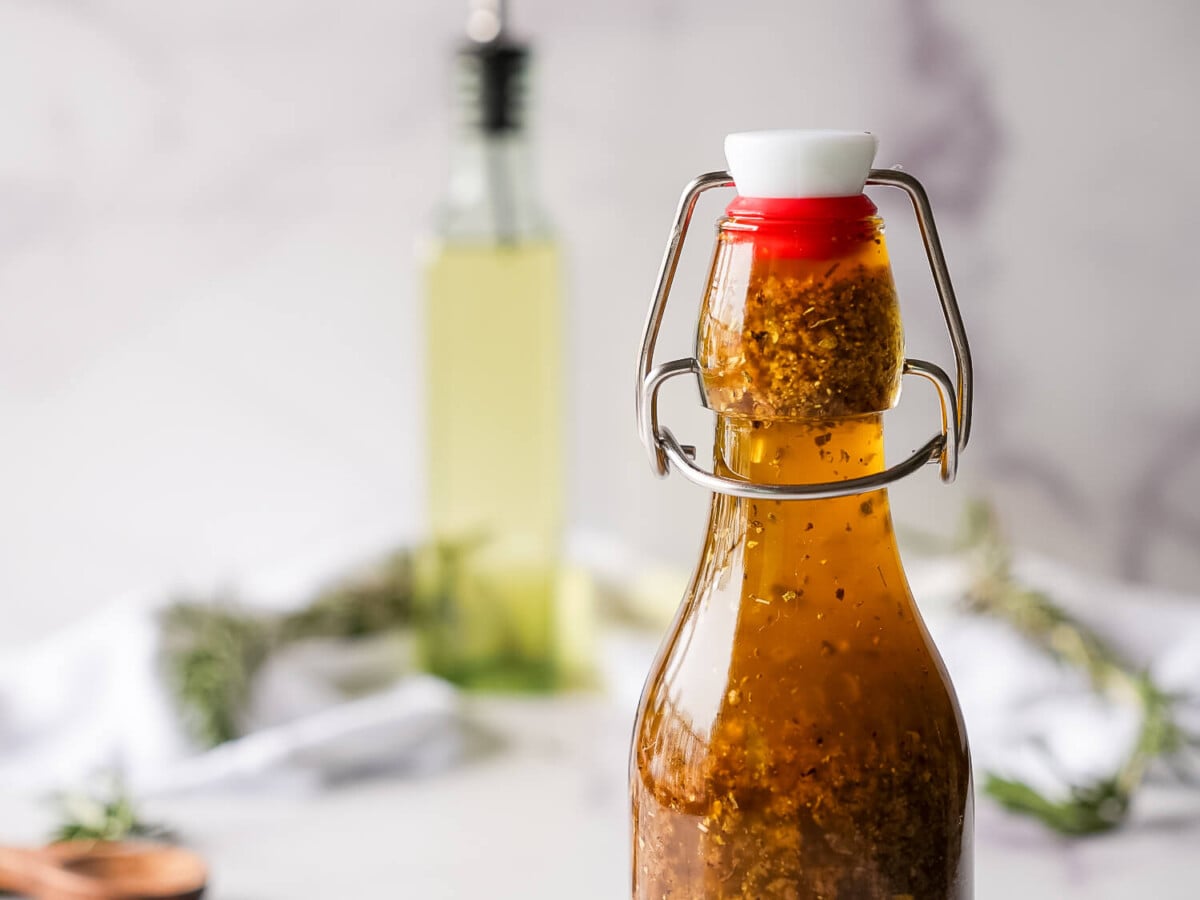 close up photo of greek salad dressing in a glass bottle in the foreground, olive oil in the background