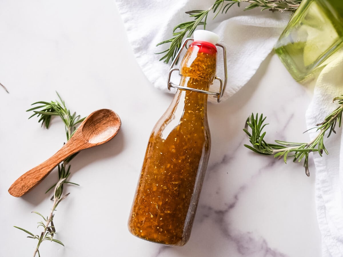 flatlay photo of greek salad dressing on a marble surface next to herbs and a wooden spoon