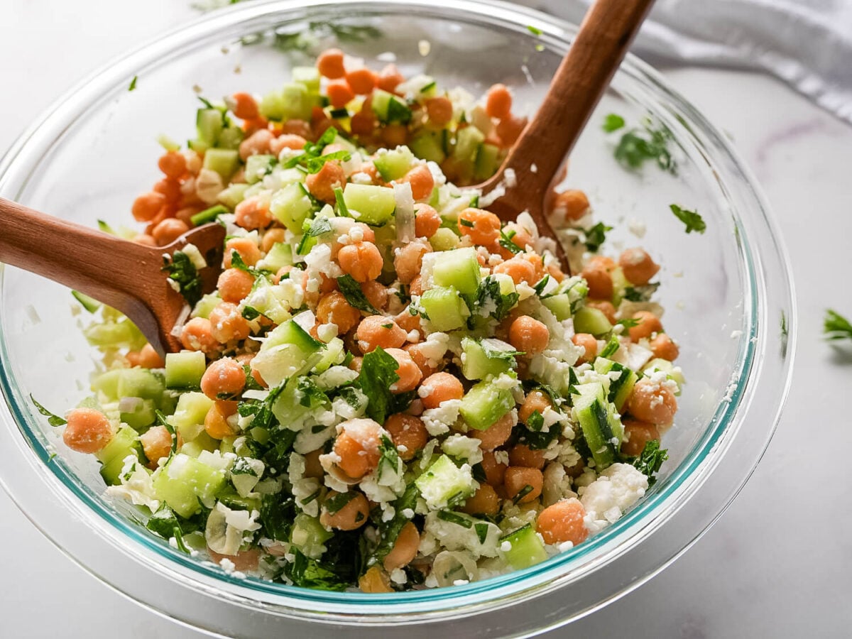 photo of a Chickpea Cucumber Salad in a glass bowl with wooden utensils on a marble surface