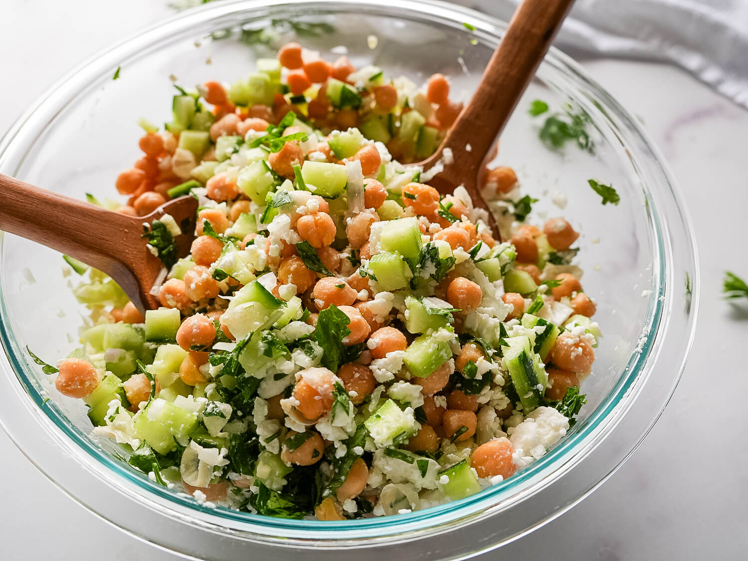 photo of a Chickpea Cucumber Salad in a glass bowl with wooden utensils on a marble surface