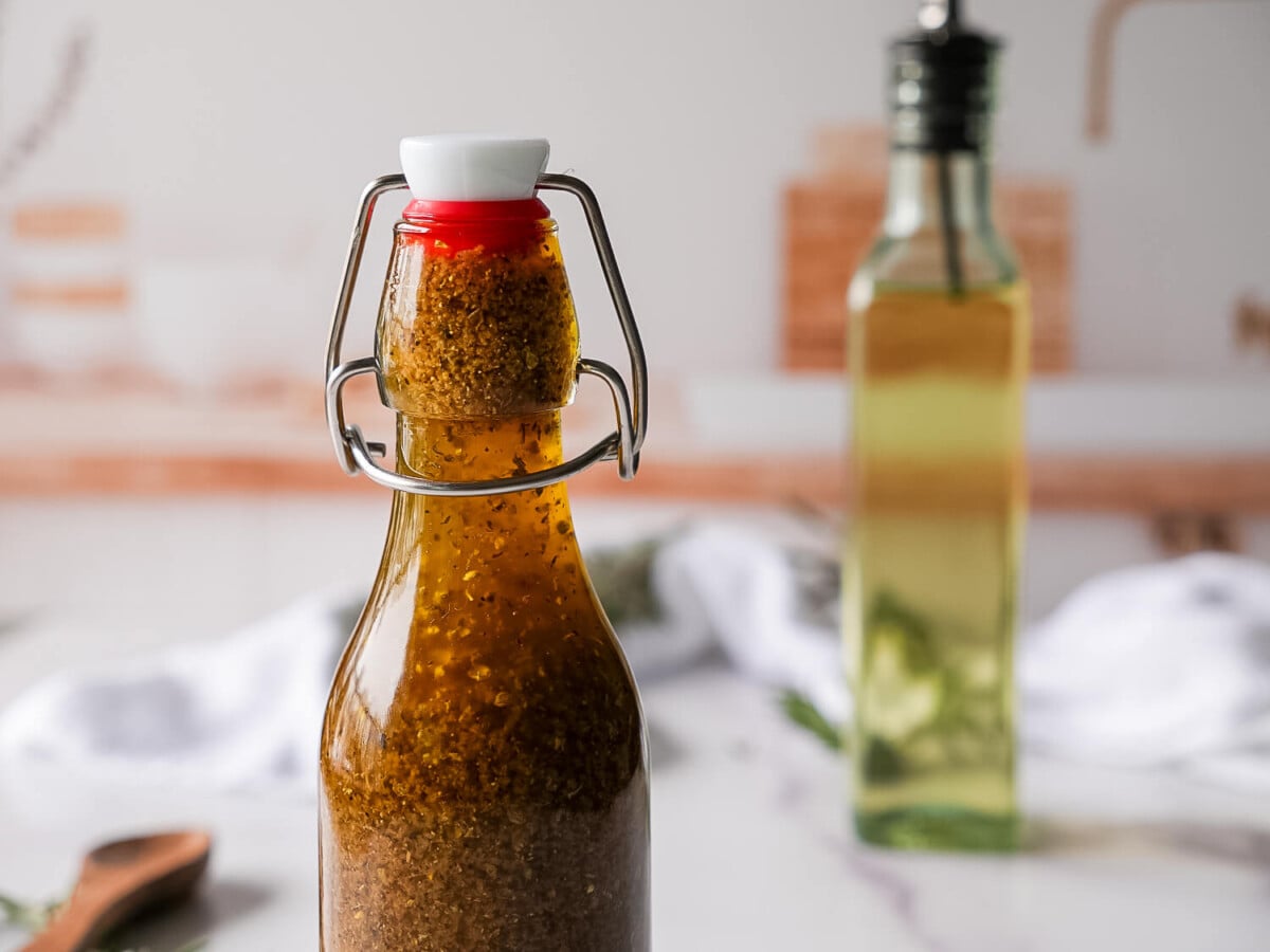 photo of greek salad dressing in a glass bottle in the foreground, olive oil in the background