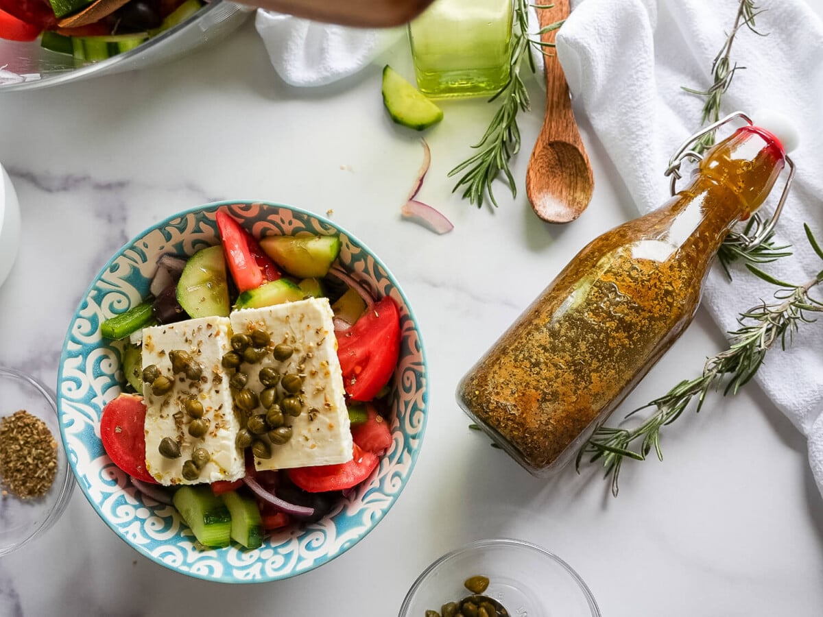 photo of greek salad in a bowl with greek salad dressing on the side in a glass bottle
