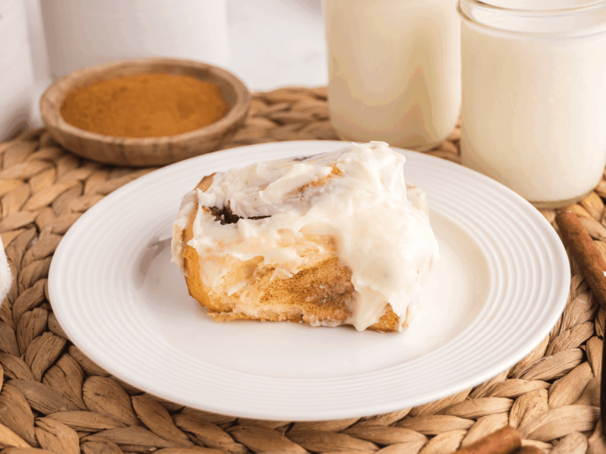 photo showing a single cinnamon roll on a white plate with two cups of milk in the background