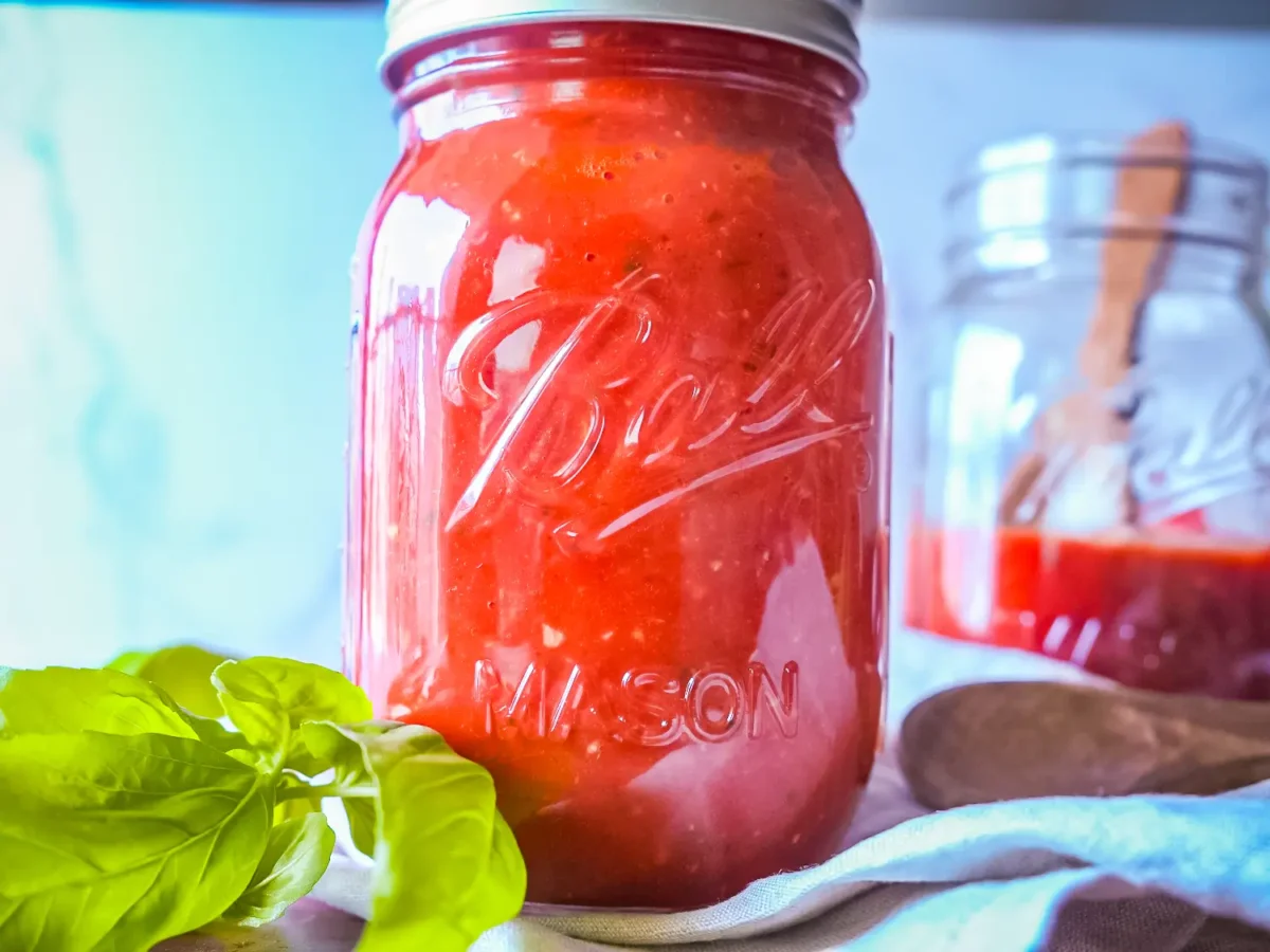 A glass mason jar filled with homemade marinara sauce sits beside fresh basil leaves, with another jar and wooden utensils softly blurred in the background
