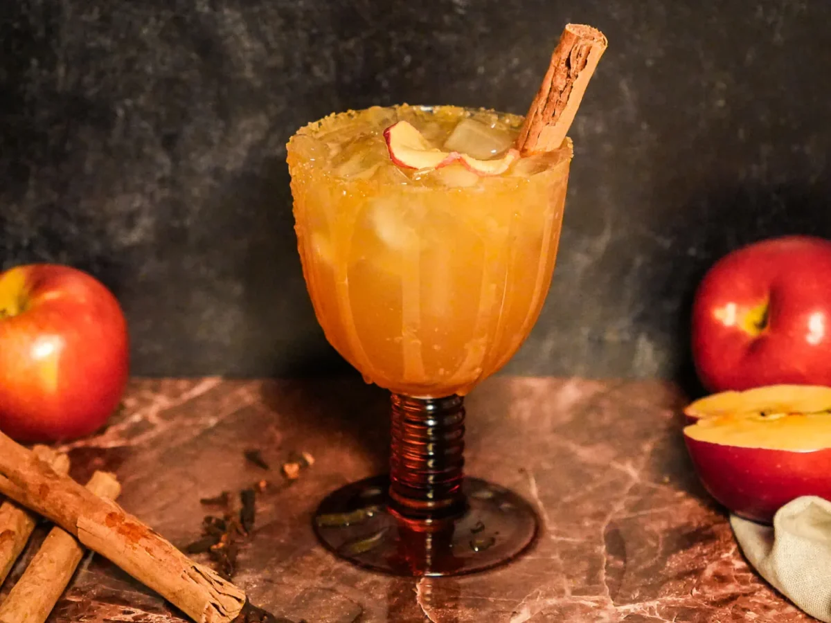 Close-up of an apple cider margarita served over ice in an amber glass with a cinnamon-sugar rim, apple garnish, and cinnamon stick