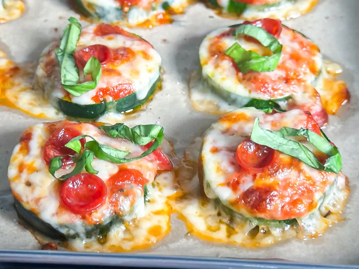 Close-up of baked zucchini pizza bites topped with marinara sauce, melted mozzarella cheese, mini pepperoni, and ribbons of fresh basil on a parchment-lined baking sheet