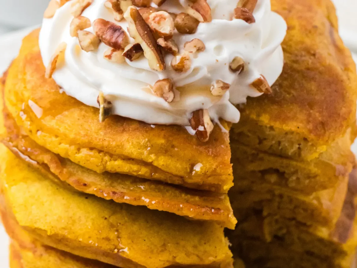 Close-up of fluffy pumpkin pancakes drizzled with maple syrup and topped with whipped cream and chopped pecans, with a cut section revealing the soft, tender interior