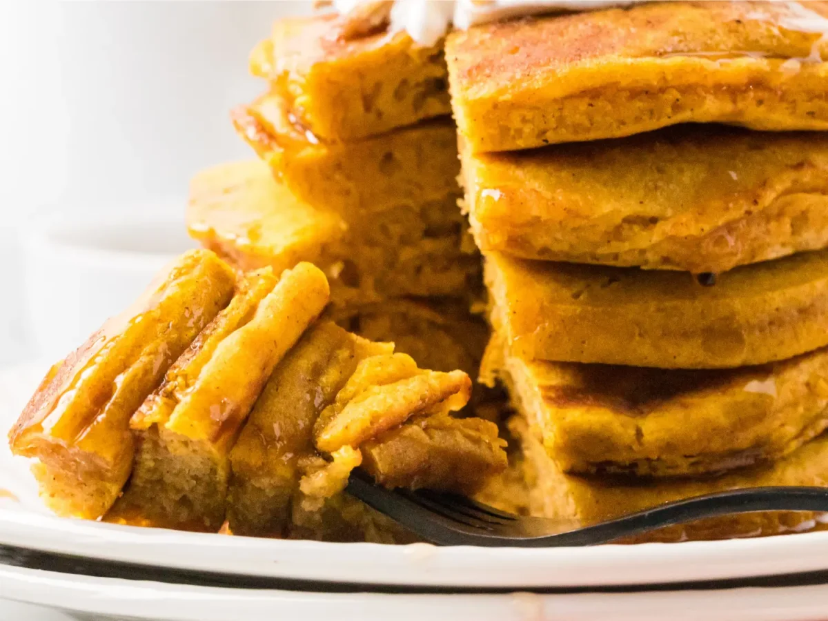 Close-up of fluffy pumpkin pancakes stacked on a white plate, drizzled with maple syrup and cut open to show their soft, tender pumpkin-spiced interior