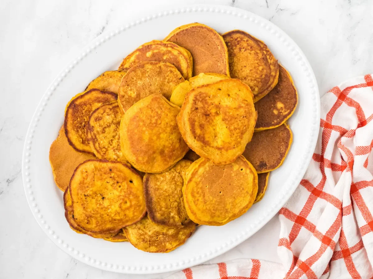 Overhead view of a large white platter filled with golden-brown pumpkin pancakes, arranged beside a red-and-white checkered kitchen towel on a light marble surface