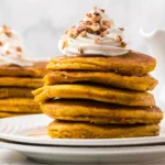 Stack of fluffy pumpkin pancakes topped with maple syrup, whipped cream, and chopped pecans, served on a white plate with more pancakes and a syrup pitcher in the background