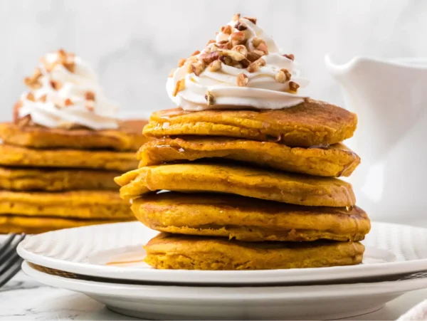 Stack of fluffy pumpkin pancakes topped with maple syrup, whipped cream, and chopped pecans, served on a white plate with more pancakes and a syrup pitcher in the background