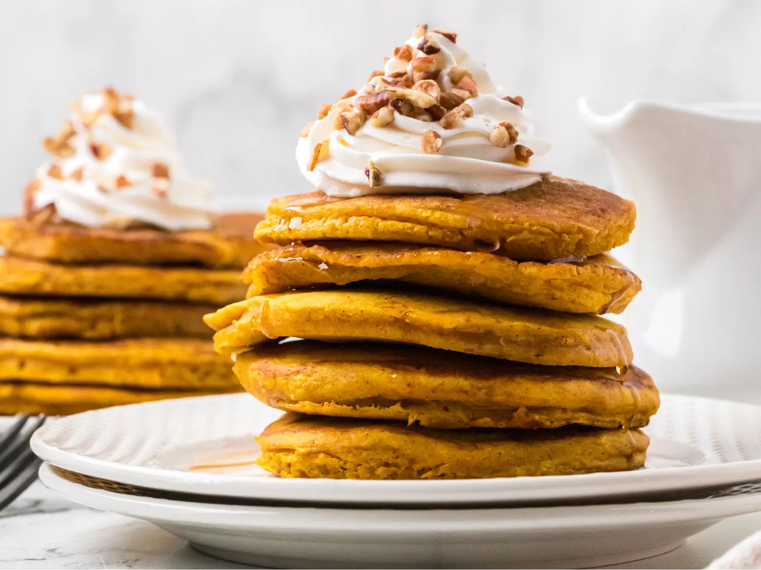 Stack of fluffy pumpkin pancakes topped with maple syrup, whipped cream, and chopped pecans, served on a white plate with more pancakes and a syrup pitcher in the background