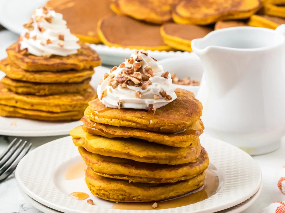 Stack of golden pumpkin pancakes topped with whipped cream, chopped pecans, and maple syrup, served on a white plate with additional pancakes and a white syrup pitcher in the background