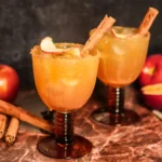 Two apple cider margaritas in amber stemmed glasses with cinnamon-sugar rims, apple garnishes, and cinnamon sticks