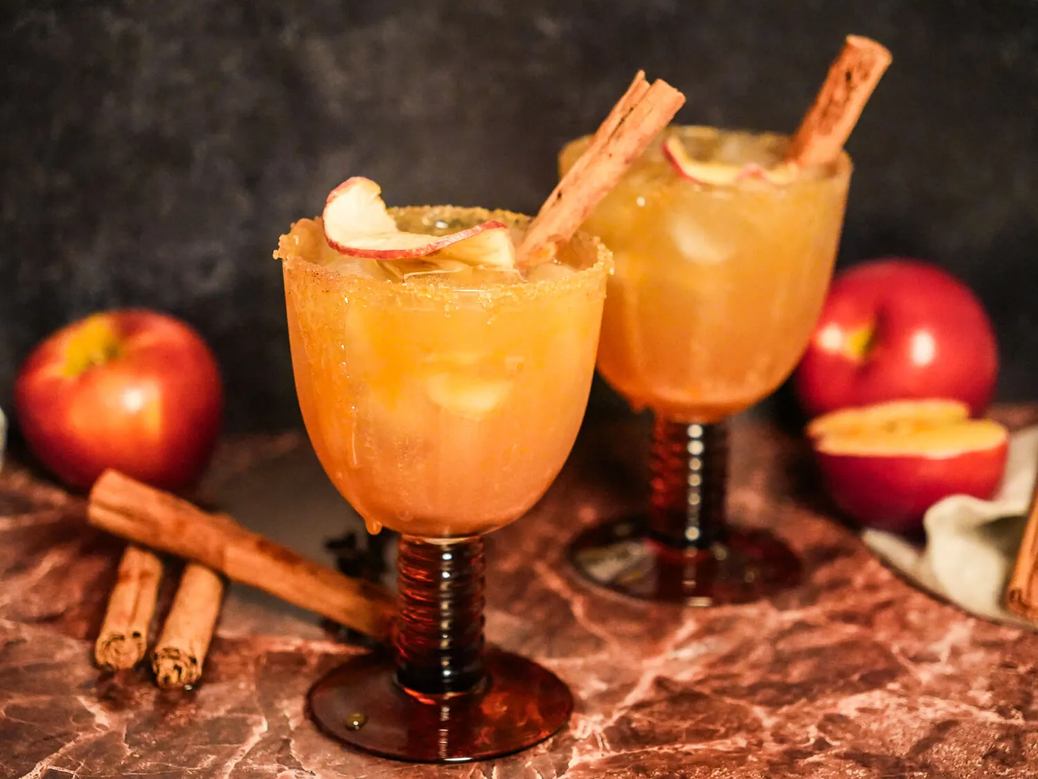 Two apple cider margaritas in amber stemmed glasses with cinnamon-sugar rims, apple garnishes, and cinnamon sticks